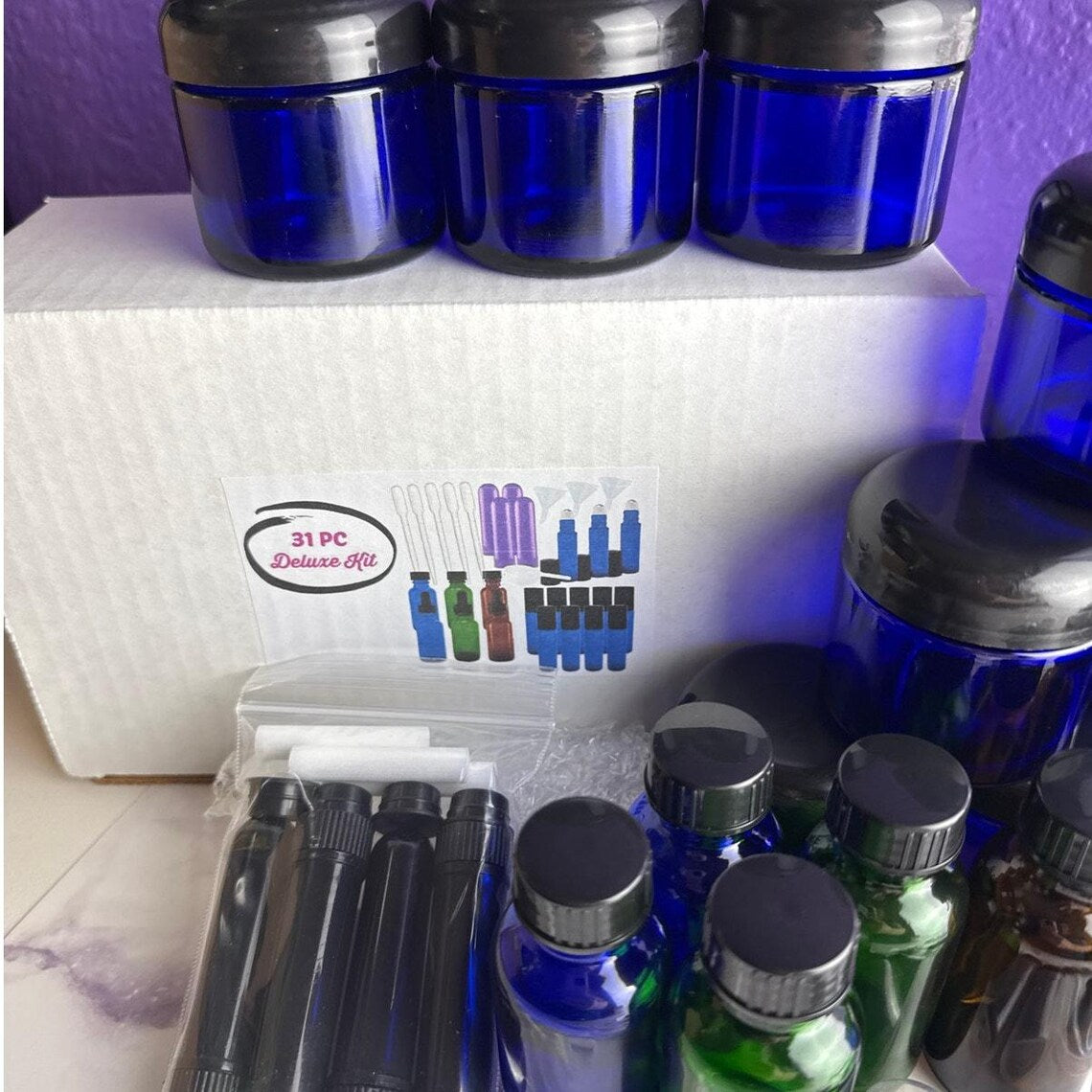 A 31-piece deluxe kit containing multiple cobalt blue glass bottles with black caps, black atomizer bottles, and a white box labeled '31 PC Deluxe Kit'.
