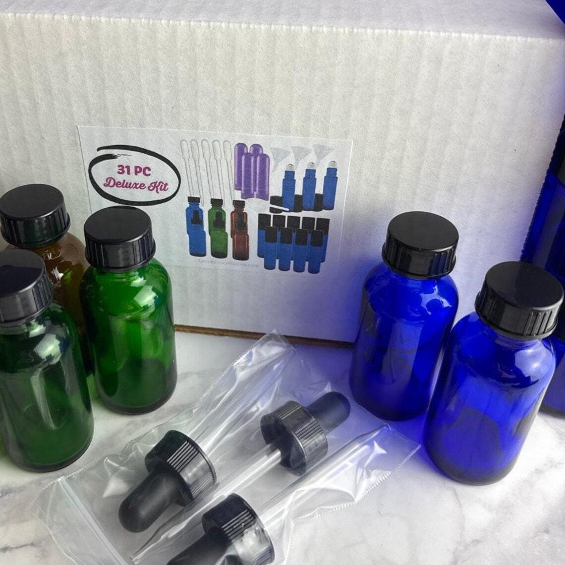 A 31-piece miniature bottle and dropper kit displayed on a white marble surface with its white cardboard packaging box in the background. The kit includes various glass bottles in green, blue, and amber, along with clear plastic dropper bottles with black rubber and plastic caps, all shown alongside the product box.