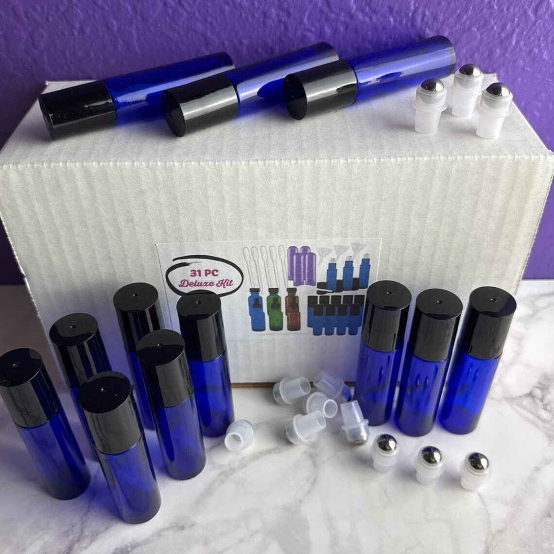 A 31-piece roll-on bottle kit displayed on a white marble surface with a purple background. The kit includes multiple cobalt blue glass roller bottles with black caps, several clear plastic roller balls, and a white cardboard box labeled '31 PC Deluxe Kit'.