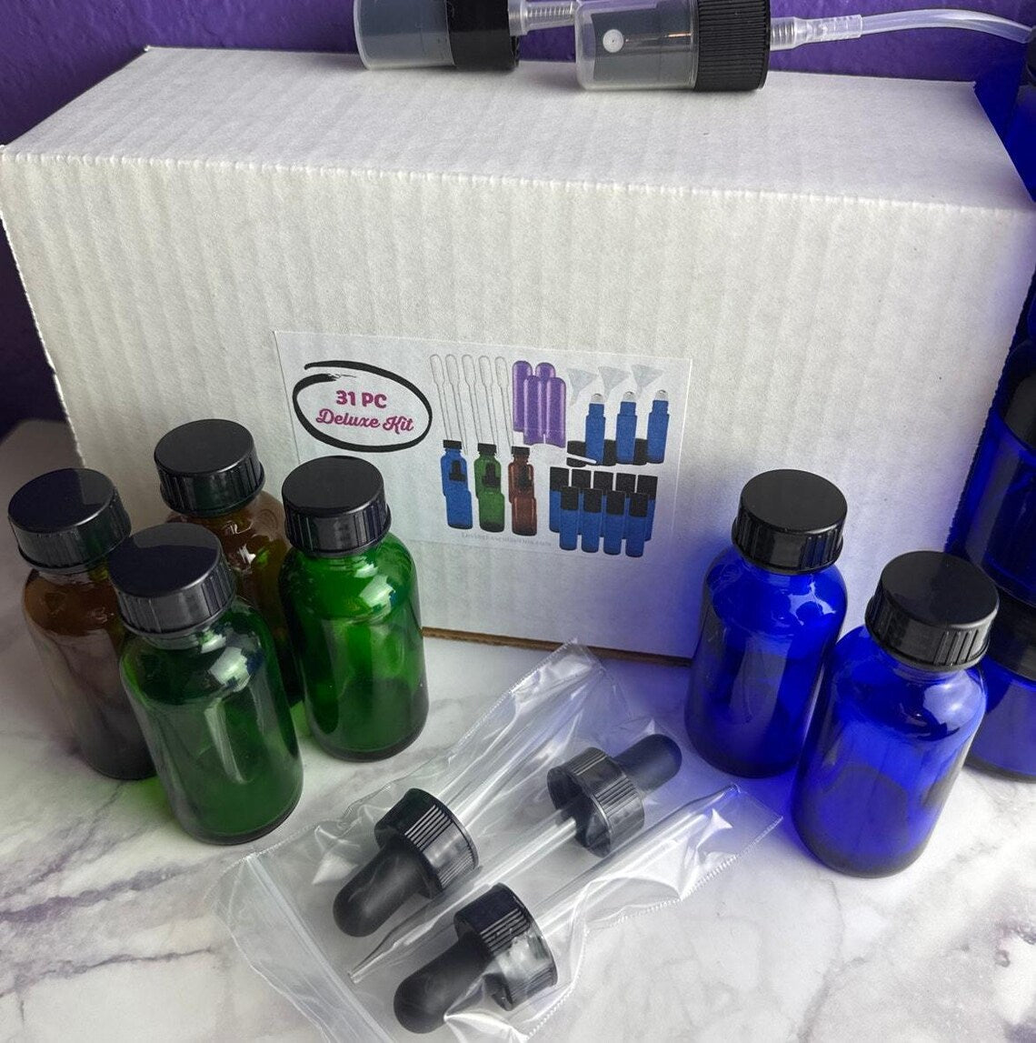 A 31-piece distill kit set displayed with its white cardboard box, featuring multiple glass distillation bottles in green, blue, and amber, along with black droppers in plastic bags, set on a white marble surface.