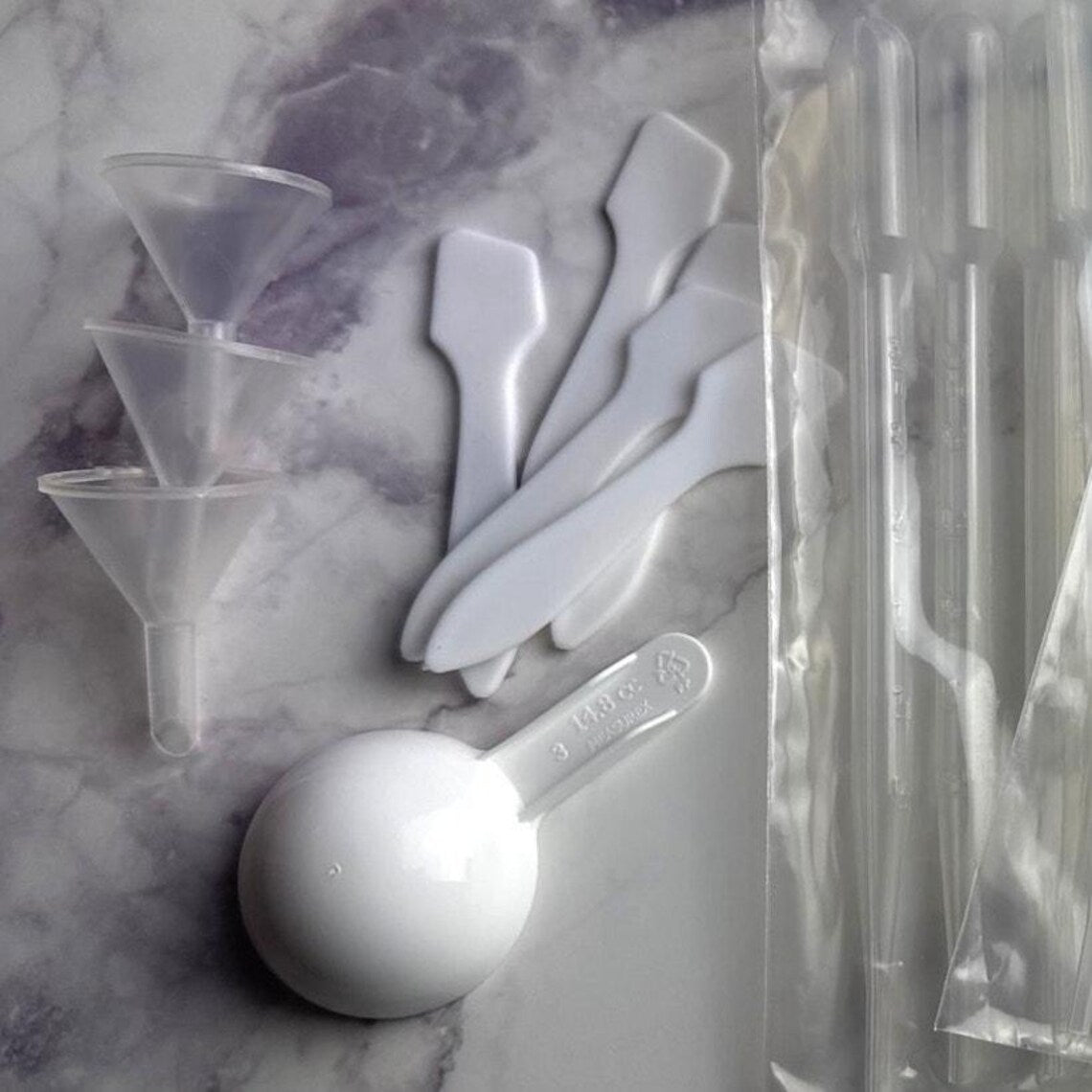 White plastic transfer tools and clear plastic funnel accessories arranged on a gray marble surface next to a sealed clear plastic bag.